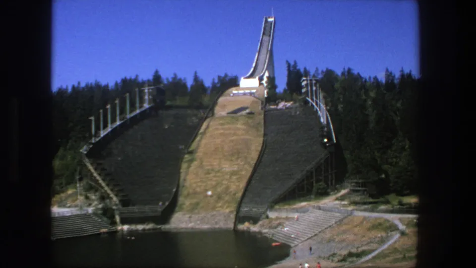 1975 home-movie frame showing the ski jump complex above water, with small visitors visible near paths at the base.