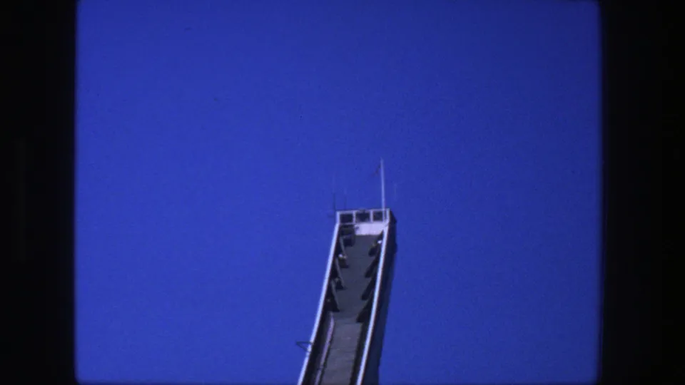1975 Stockholm 8mm frame showing the upper section of a white ski jump ramp rising into a clear blue sky.