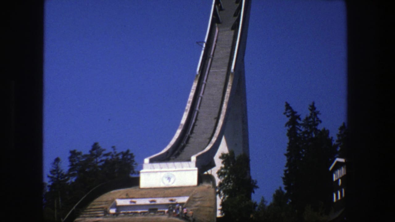 Tall ski jump ramp rising above a wooded hillside under a blue sky.