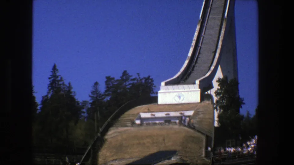 1975 Stockholm 8mm frame showing the ski jump support, wooded backdrop, grassy hill, and a small building below the ramp.