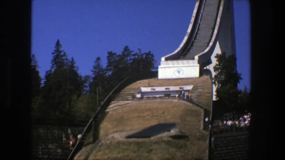 1975 Stockholm 8mm frame showing the ski jump base, grassy landing area, surrounding trees, and distant spectators along the right side.