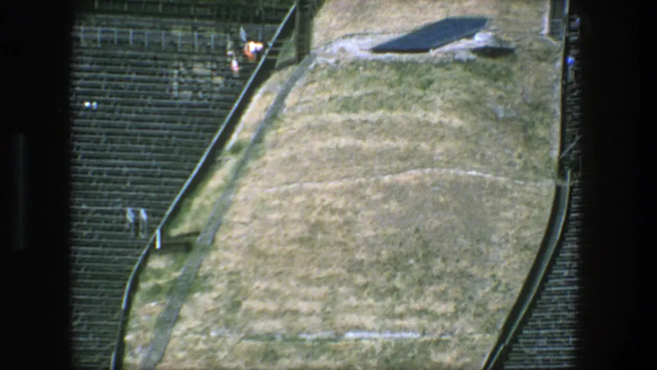 1975 Stockholm 8mm frame showing tan open ground, dark seating rows, railings, and a sloped outdoor edge.