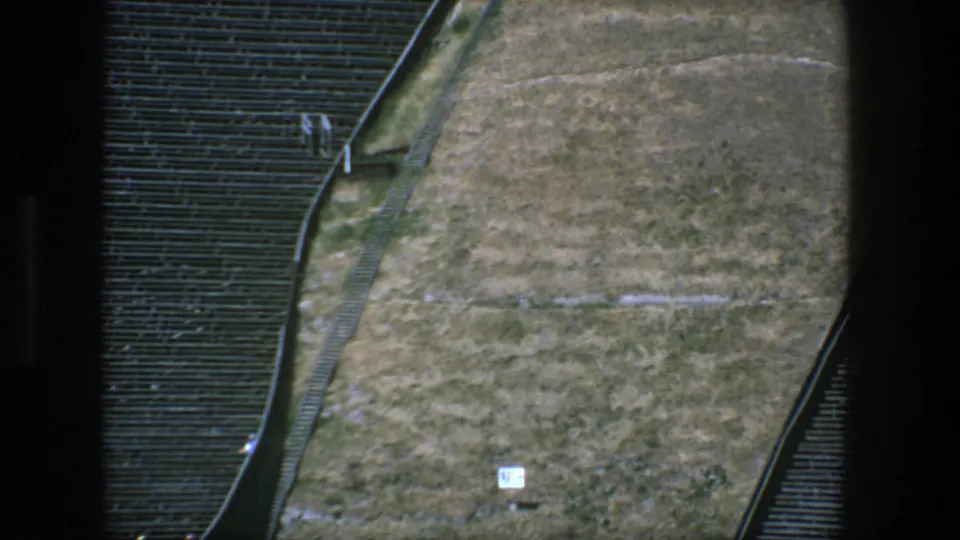 1975 Stockholm 8mm frame showing a tan field-like area, small sign, seating rows, and curved boundary rail.