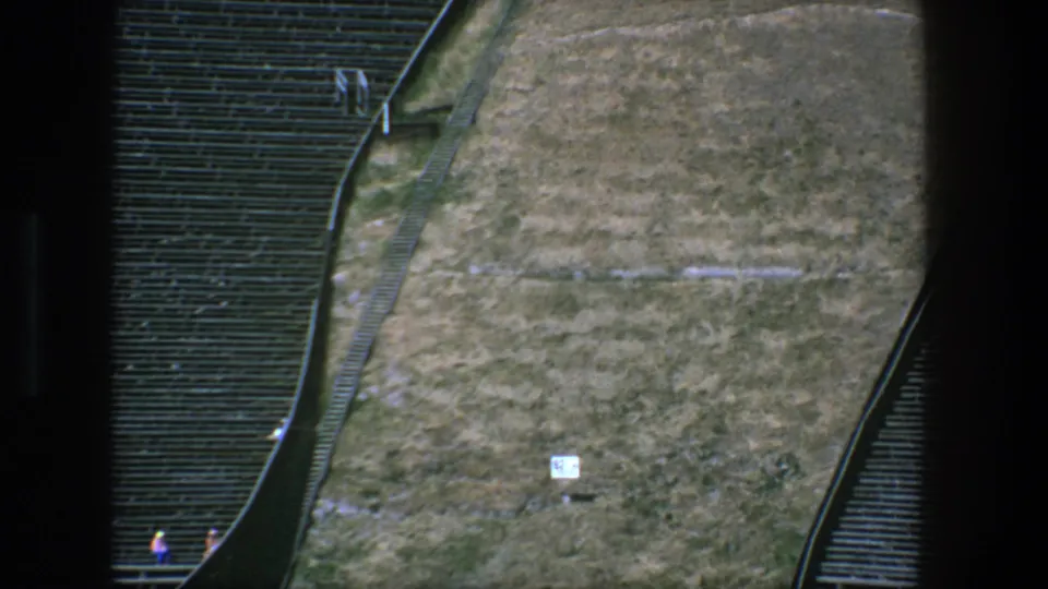 1975 Stockholm 8mm frame showing tan ground, a small sign, dark seating rows, railings, and tiny distant figures.