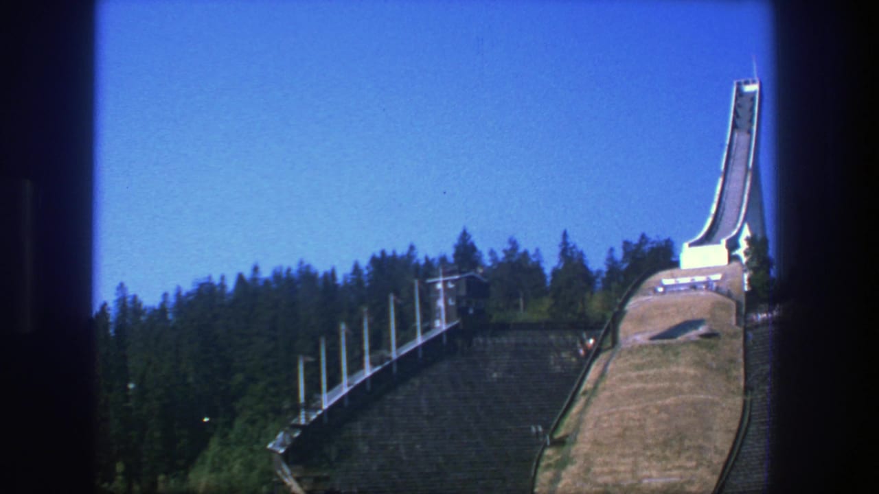 Ski-jump ramp rising from a grassy hillside beside dark pine forest under blue sky.