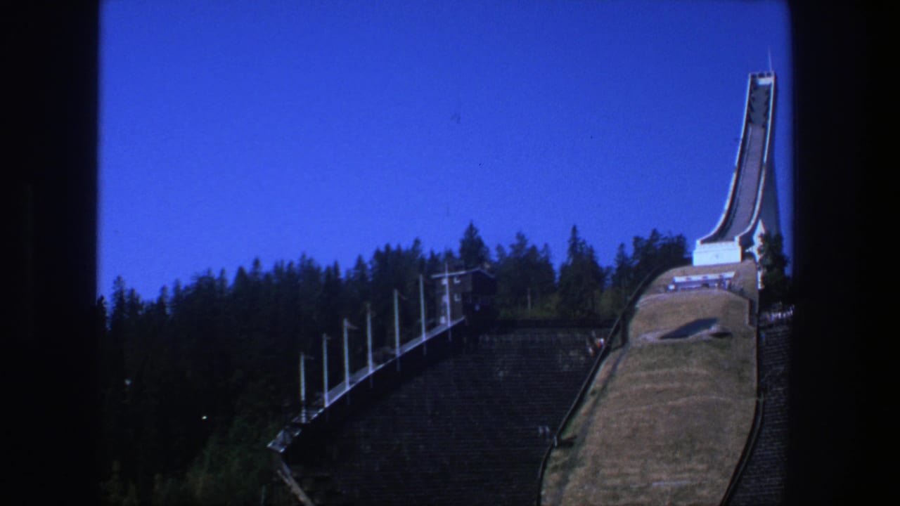 Wide view of a ski-jump structure with forested slope and clear blue sky.