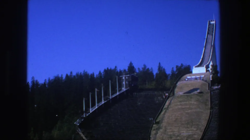 1975 Stockholm view of the ski-jump ramp and tower, framed against pine trees and a saturated blue sky.
