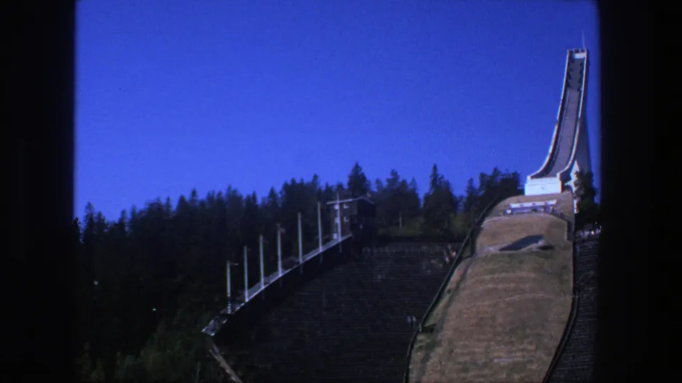 1975 Stockholm 8mm frame of a ski-jump ramp viewed from below, with pine trees and hillside details visible.