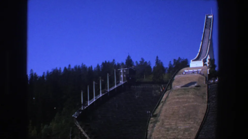 1975 Stockholm frame showing the ski-jump tower, ramp, grassy slope, and surrounding pine forest in daylight.