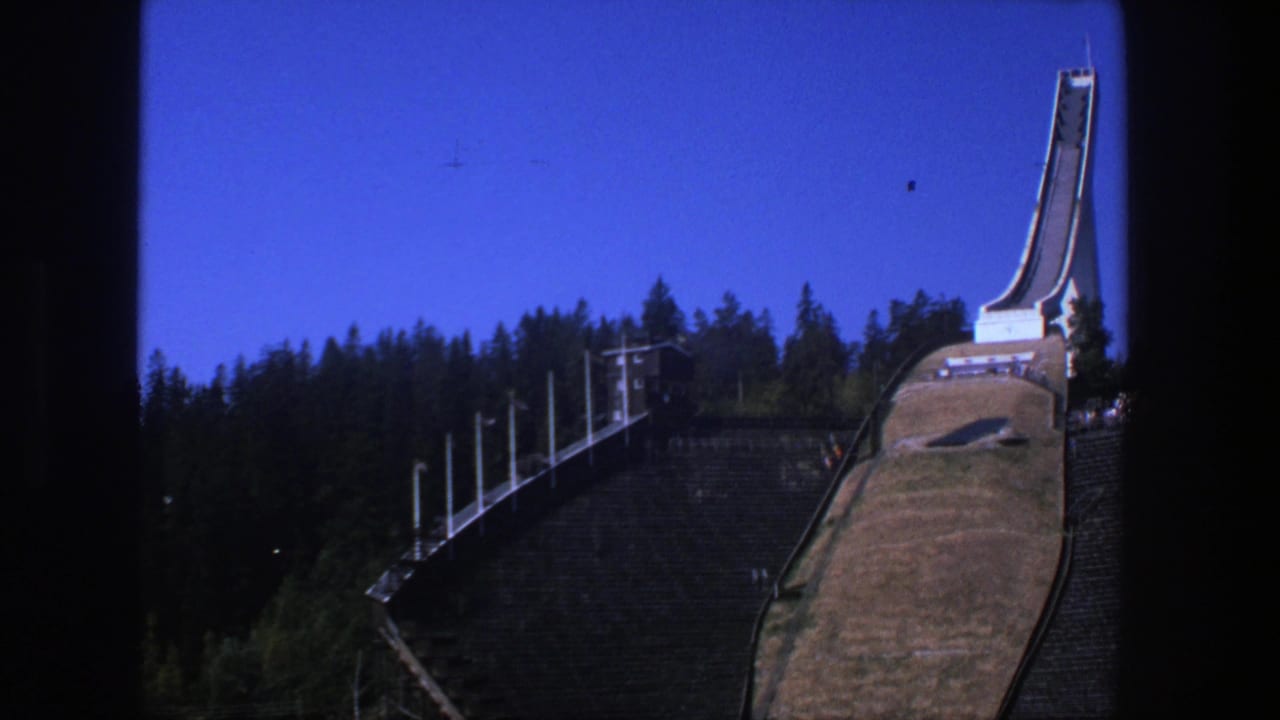 1970s 8mm travel frame showing a tall ski jump above trees and empty spectator stands, with dark film edges visible.
