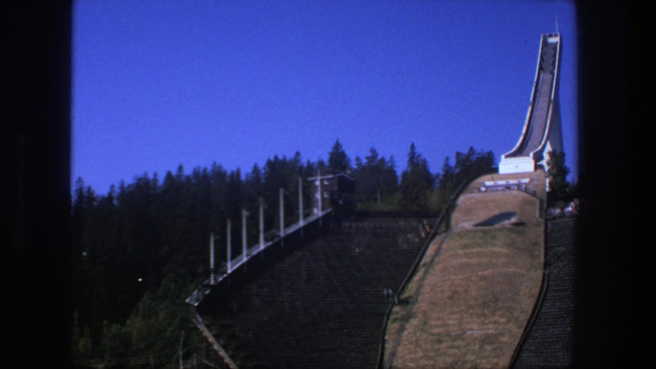 1970s 8mm frame of the ski jump landmark from a visitor viewpoint, with forest, stands, and strong color saturation.