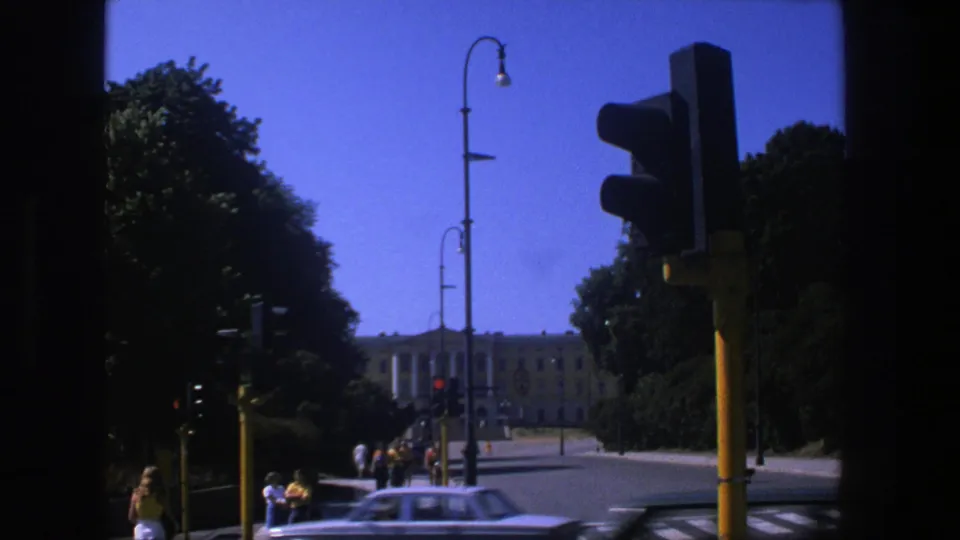 1970s 8mm Stockholm street scene with pedestrians near yellow traffic lights, passing cars, trees, lamps, and a distant civic building.
