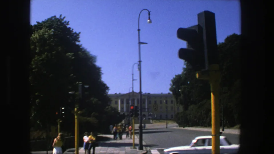 1970s 8mm Stockholm street frame showing a traffic signal in the foreground, pedestrians on the sidewalk, cars, trees, and a distant building.