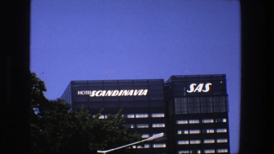 1975 Stockholm 8mm frame showing Hotel Scandinavia and SAS signage on a modern building under a clear blue sky.