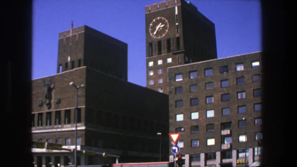 1975 Stockholm street view of a brick civic building with a clock tower, traffic lights, and road signs under a blue sky.