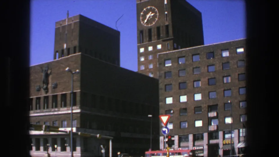 1975 Stockholm street scene with a brick public building, clock tower, traffic lights, signs, and a red bus passing below.