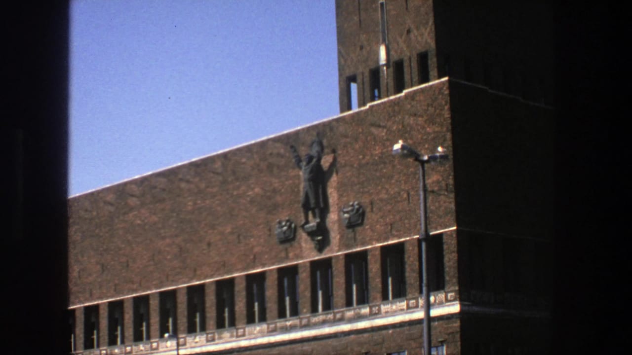 1975 Stockholm exterior frame with a raised-arm wall figure on a brown brick building, flanking emblems, narrow windows and clear sky.