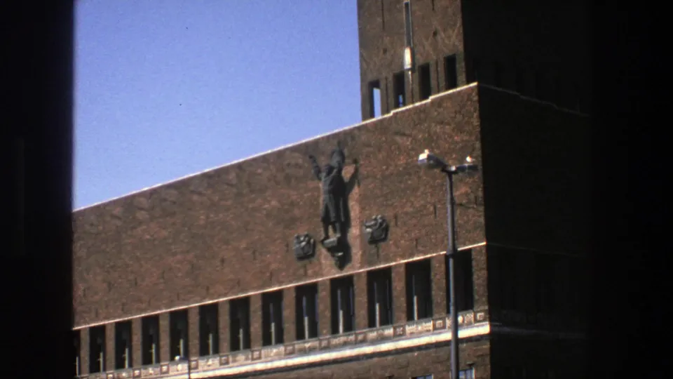 1975 Stockholm exterior frame with a raised-arm wall figure on a brown brick building, flanking emblems, narrow windows and clear sky.