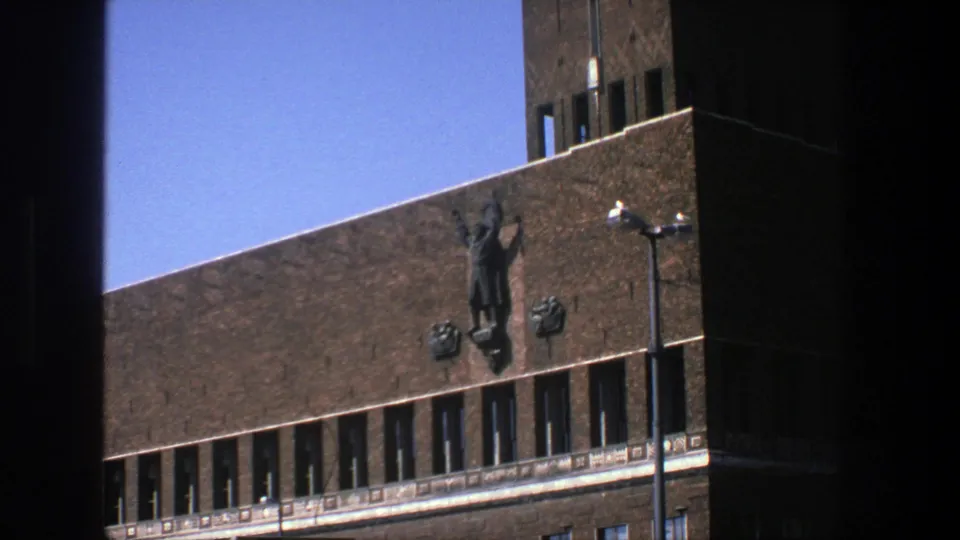 1975 Stockholm 8mm view of an angled brick building facade, showing a mounted figure sculpture above narrow windows and a foreground streetlamp.
