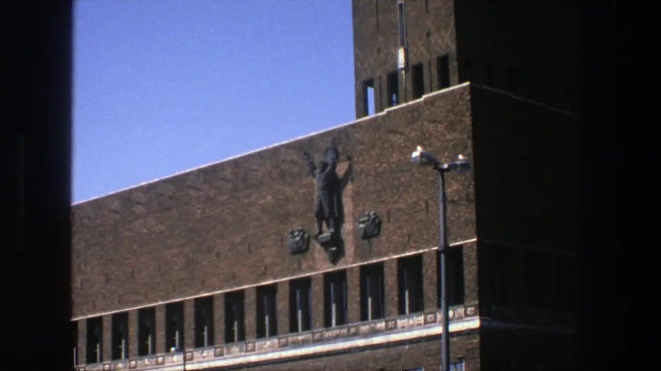 1975 Stockholm frame of a brick building corner with a raised-arm facade sculpture, emblems, window band, streetlamp and blue sky.