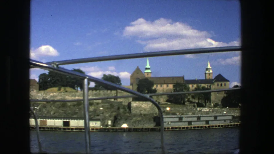 1975 boat-view footage in Stockholm showing railings, harbor water, rocky stonework, trees, and towered architecture.