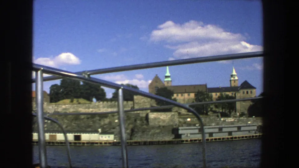 1975 8mm Stockholm boat-view frame with metal railings, rocky shore, stone walls, trees, and spired historic architecture across the water.