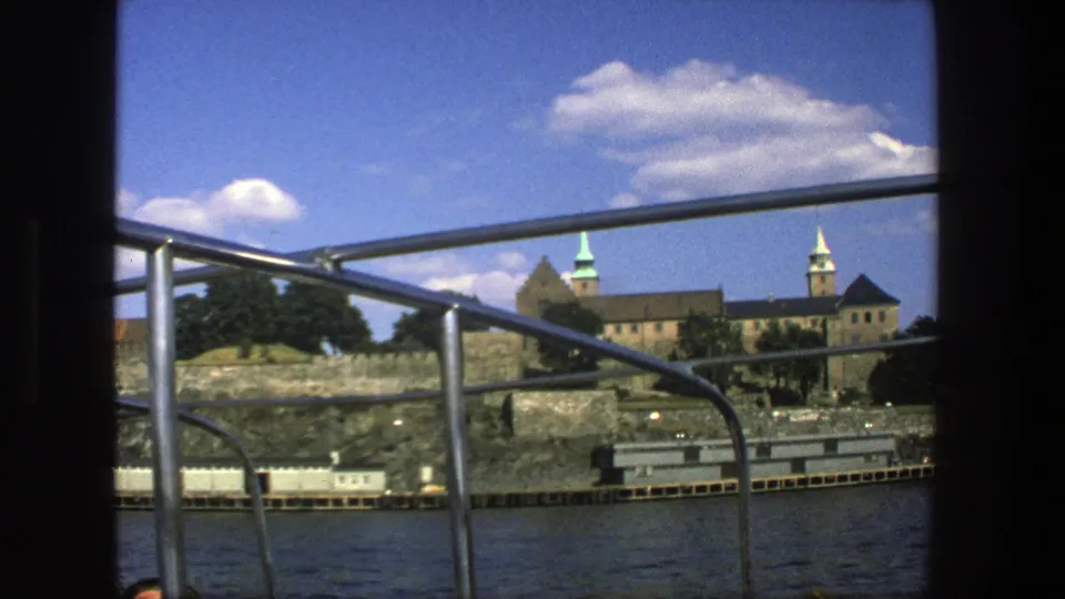 Stockholm waterfront architecture appears behind metal boat railings, with stone walls, trees, water, and bright clouds in a 1975 travel-film frame.