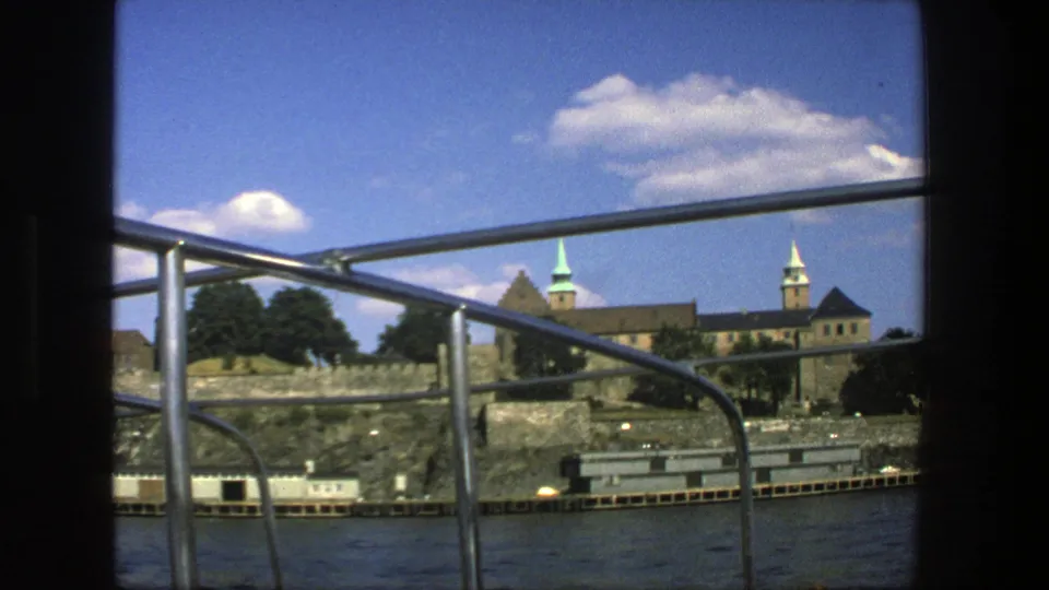 A 1975 Stockholm boat-ride view shows railings in the foreground, choppy water, rocky shore, trees, and spired historic buildings in the distance.