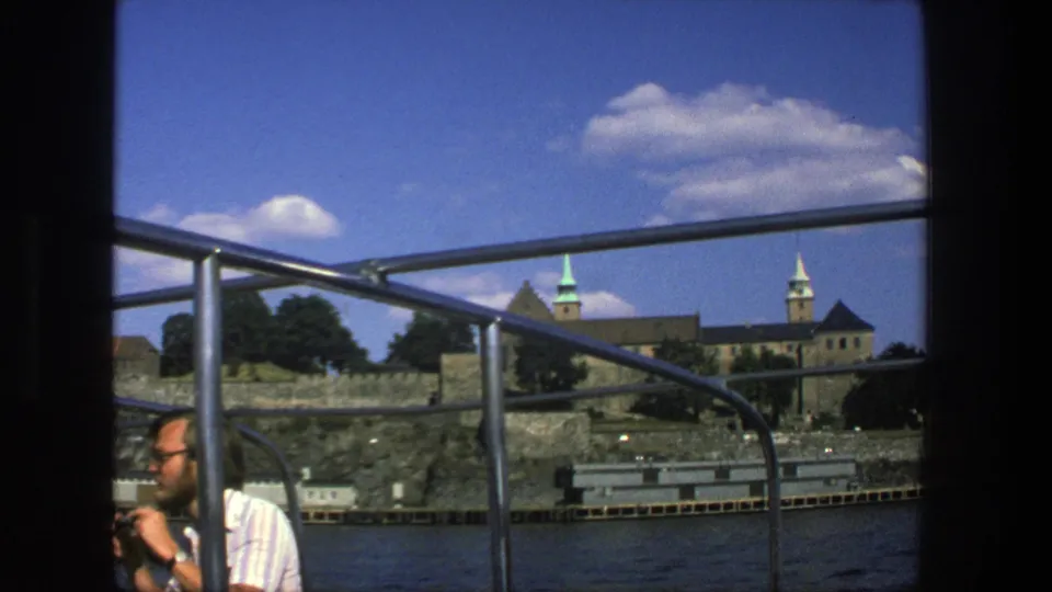In this 1975 Stockholm boat scene, a man in a striped shirt is partly visible beside the railing as spired waterfront buildings pass behind him.