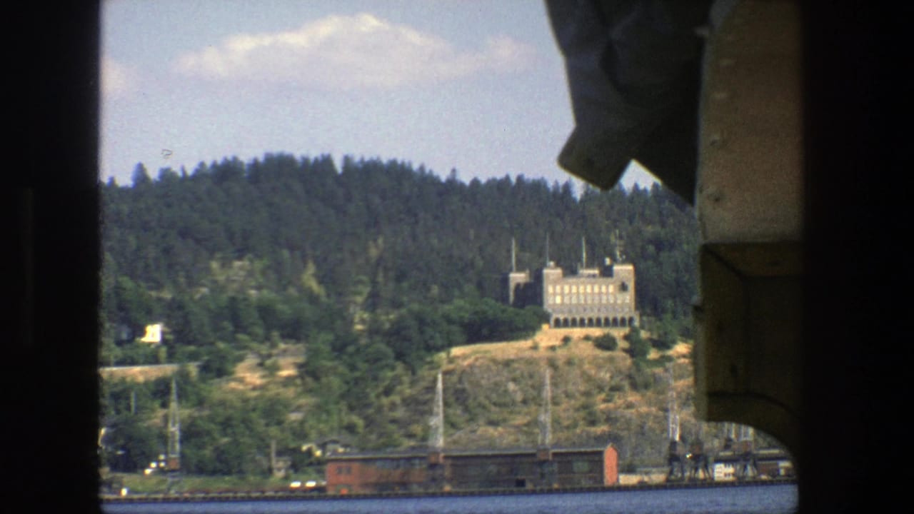 Boat-framed view across water to a wooded hill with a pale building and red rail car.
