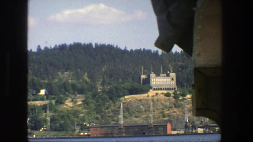 1975 Stockholm boat view across blue water toward a wooded hillside, pale spired building, and red shoreline rail car.