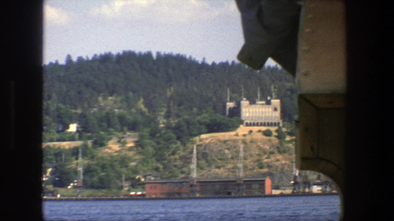 1975 Stockholm shoreline seen from a moving boat, with a wooded hill, pale building, rail car, and dark vessel framing.