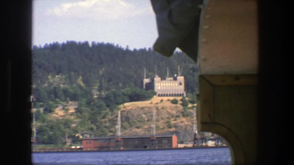 Soft 8mm frame from Stockholm in 1975 showing water, a wooded hillside, pale building, and red rail car from a boat.