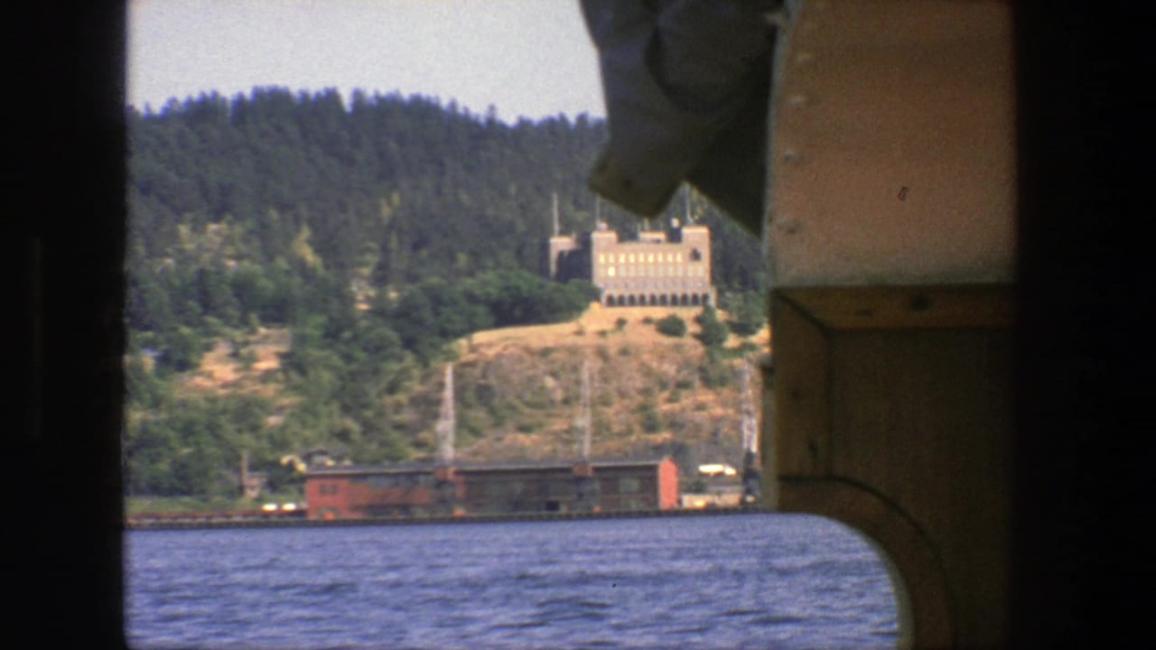 Boat-framed view of blue water, wooded hillside, tan building, and waterfront rail cars.