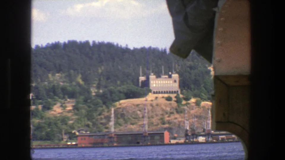 1975 Stockholm harbor view with boat framing, blue water, waterfront rail cars, and a tan building on a wooded hillside.