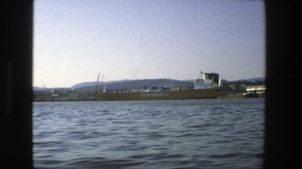 1975 Stockholm harbor scene from a boat, showing a cargo vessel on the water with cranes and distant shoreline beyond.