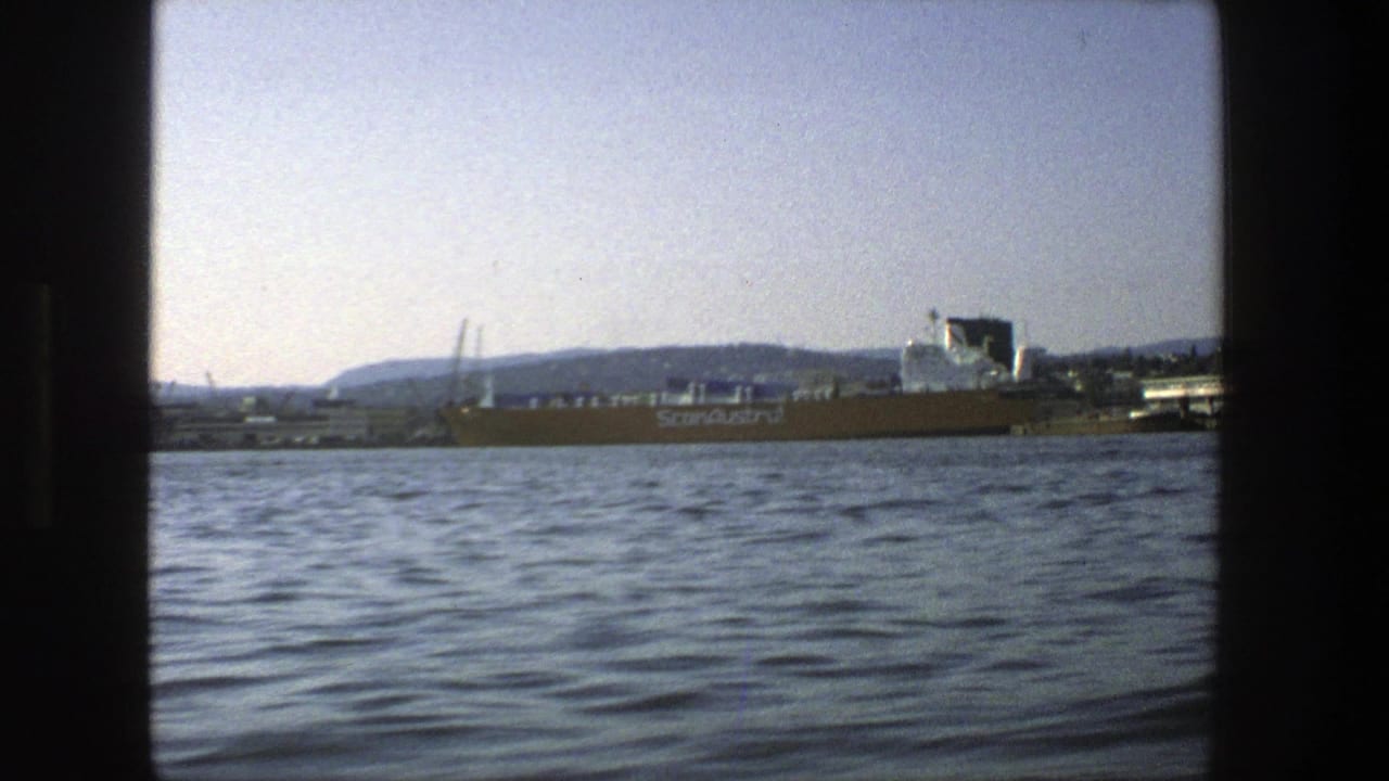 Cargo ship on Stockholm harbor water with distant hills and cranes.