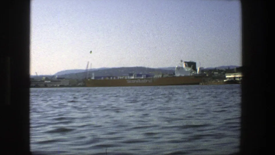 1975 home-movie frame of Stockholm harbor with a ScanAustral ship beside port structures and low hills beyond the water.