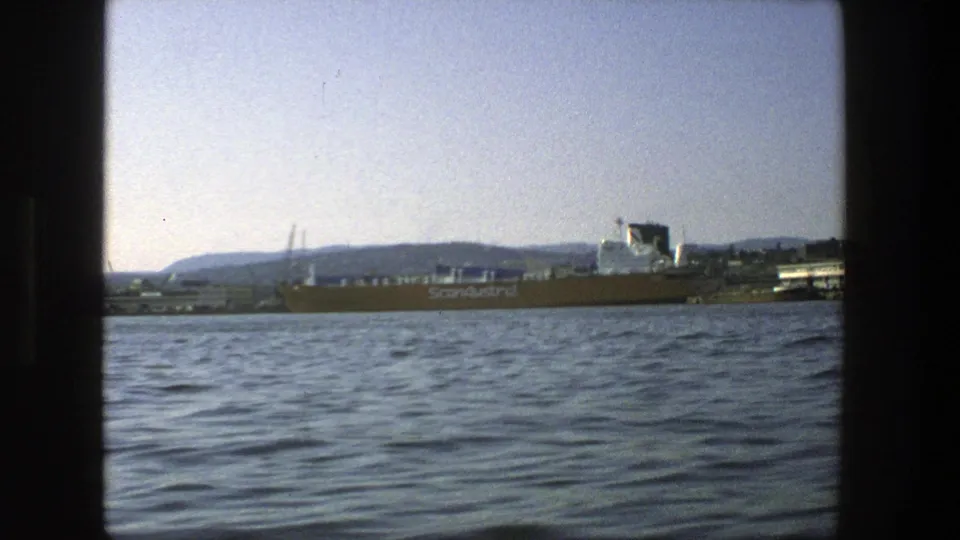 Stockholm harbor in 1975 with a ScanAustral cargo ship, industrial shoreline, rippled water, and distant hills framed by 8mm gate edges.