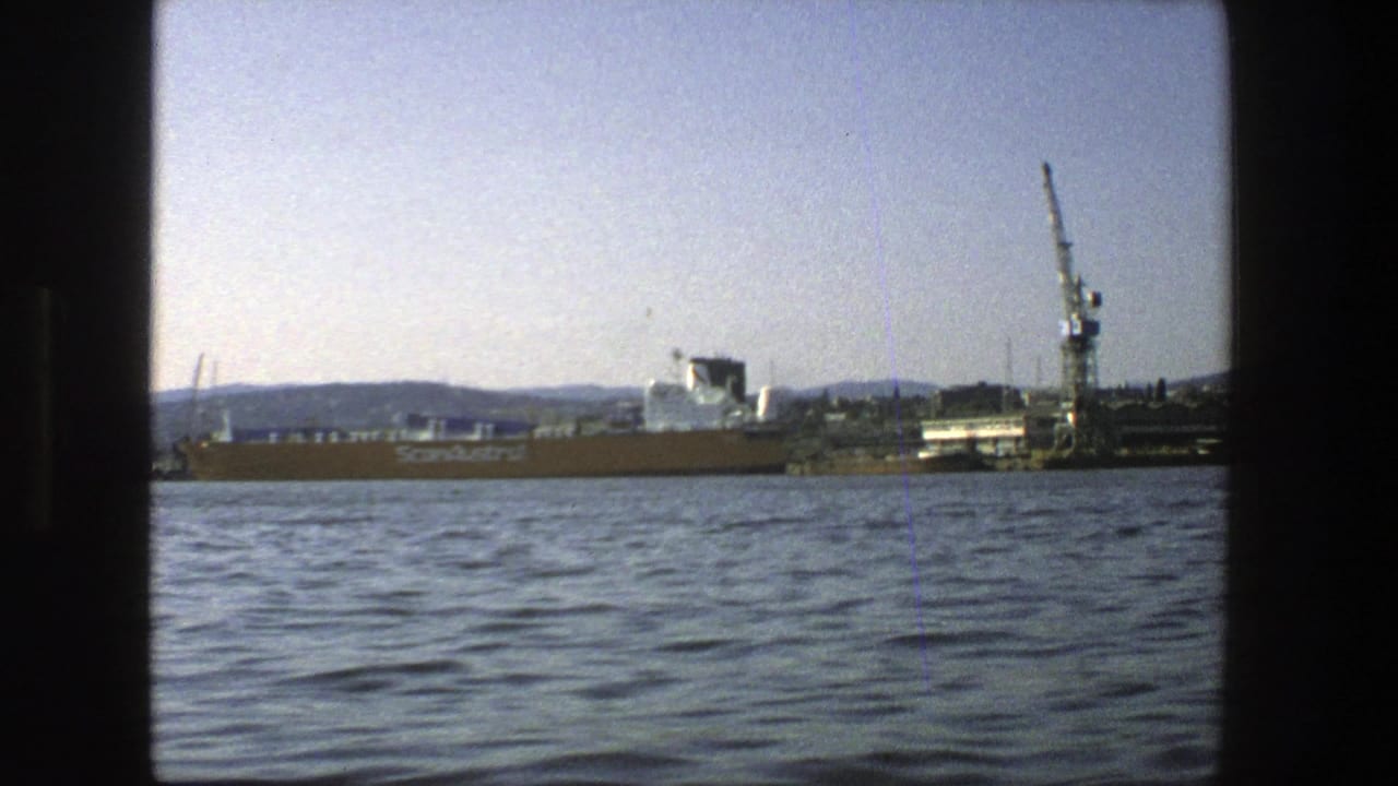Harbor water in foreground with cargo ship, dock buildings, and tall crane beyond.