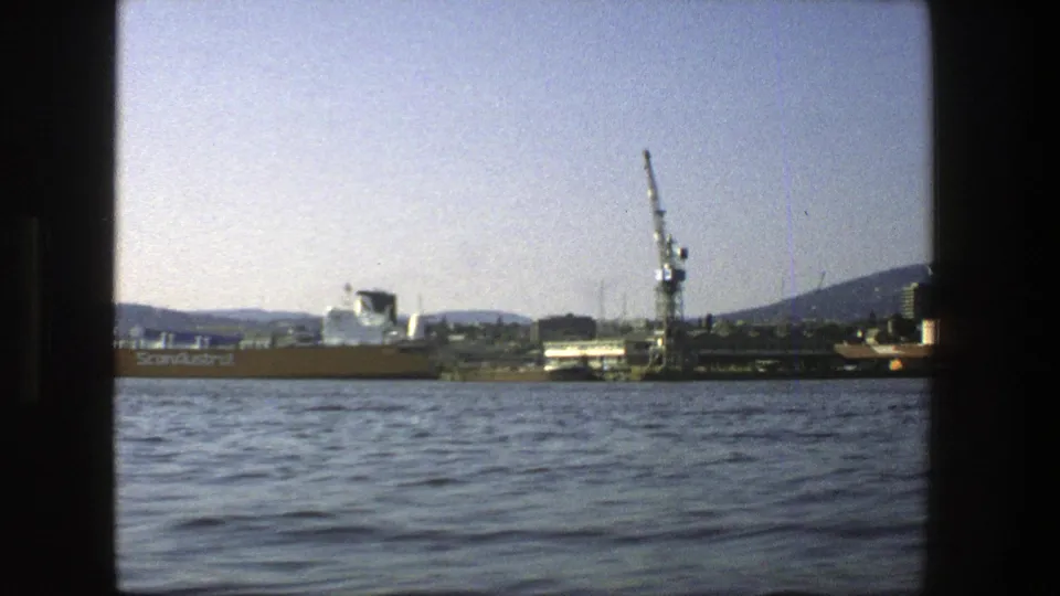Late shot of the 1975 Stockholm harbor view, with the cargo ship partly left of frame and a dock crane beside low buildings.