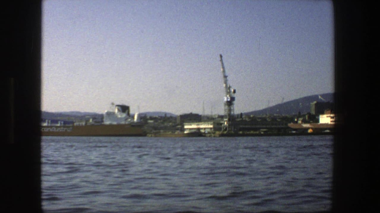 Waterfront seen from water with cranes, low buildings, distant hills, and dark film-frame borders.