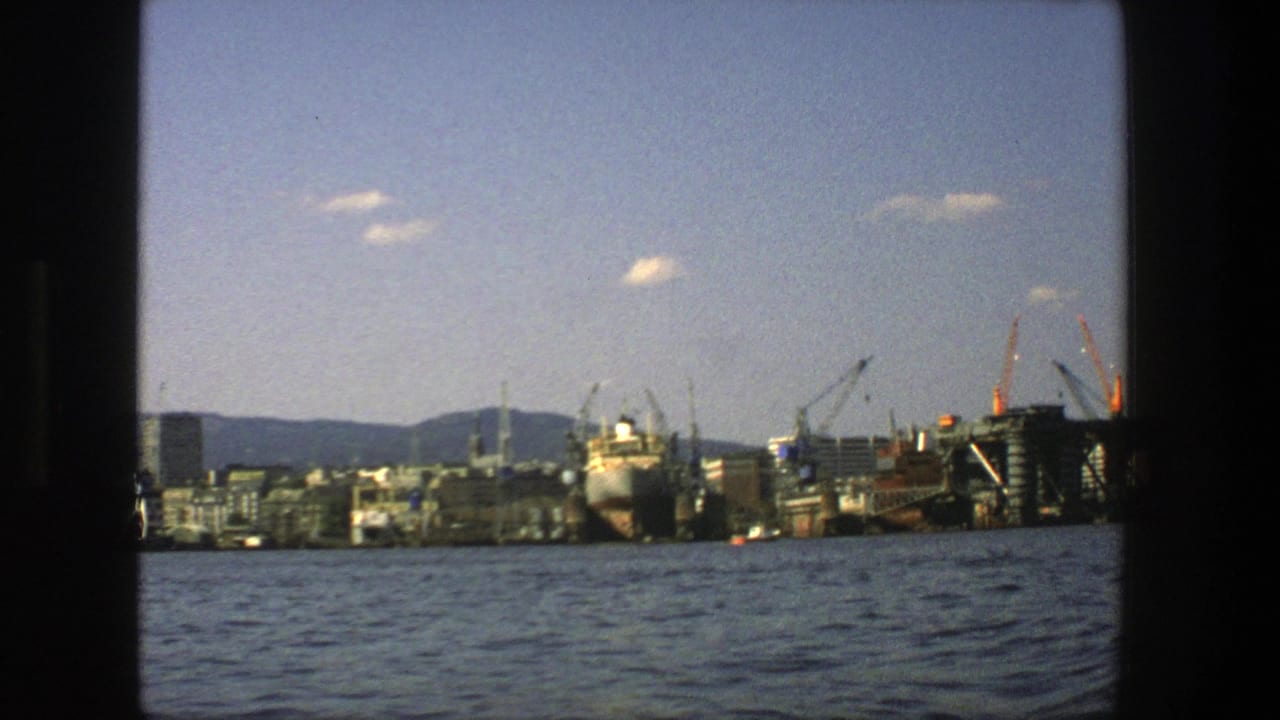 Industrial waterfront with cranes, a moored ship, buildings, hills, and open sky beyond the water.