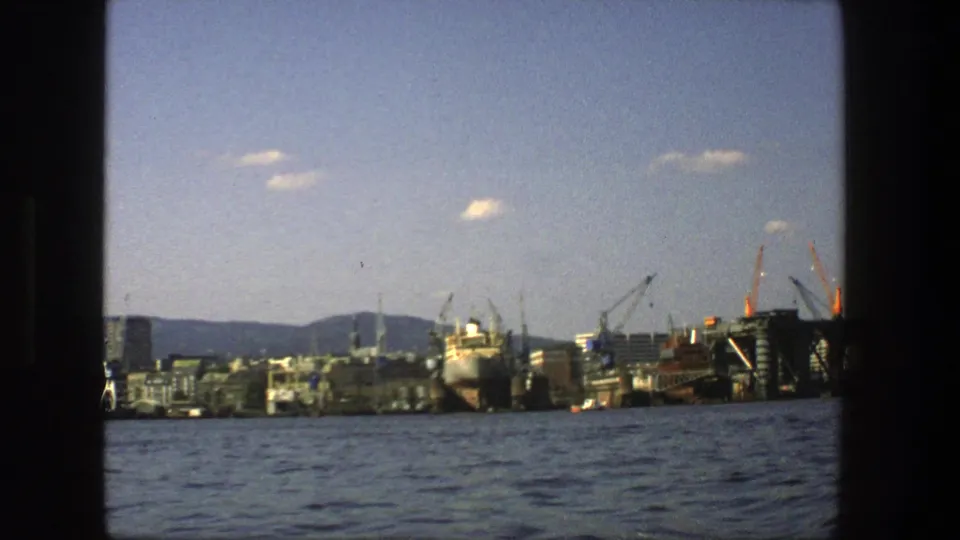 1975 Stockholm waterfront footage showing a large docked ship, multiple cranes, shoreline buildings, hills in the distance, and the harbor surface in front.