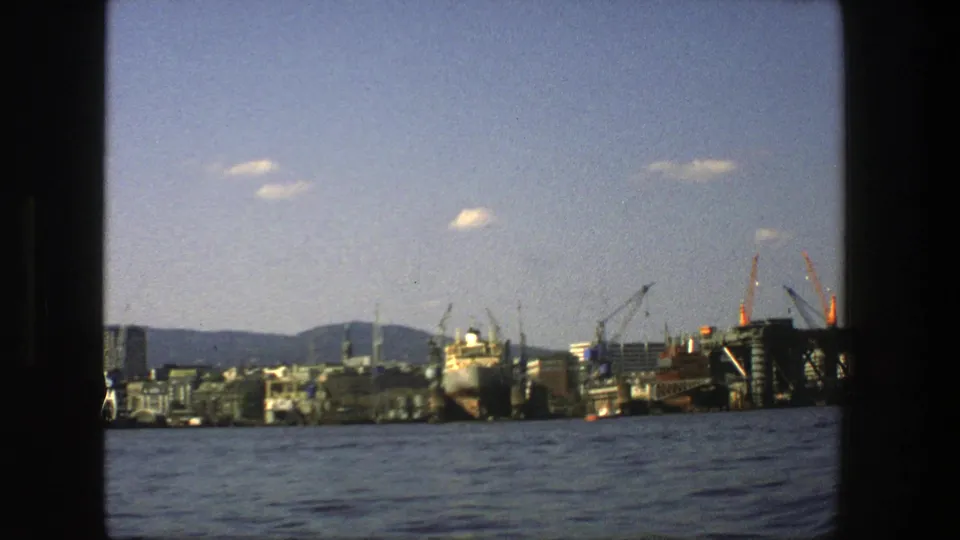 Late shot of the 1975 Stockholm harbor view, with cranes, a docked ship, waterfront construction, distant hills, and broad blue water in the foreground.