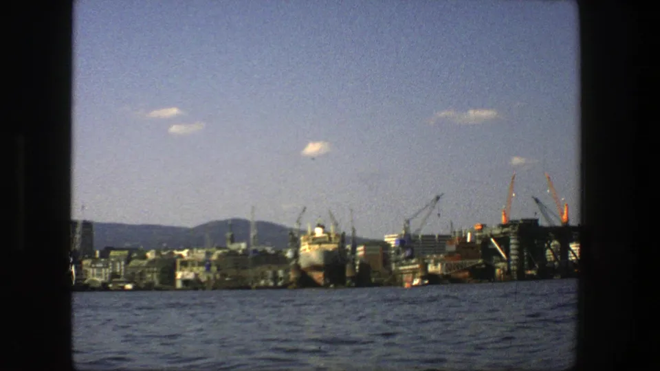 1975 Stockholm harbor view from the water, with a large vessel, industrial cranes, waterfront buildings, distant hills, and dark frame edges.