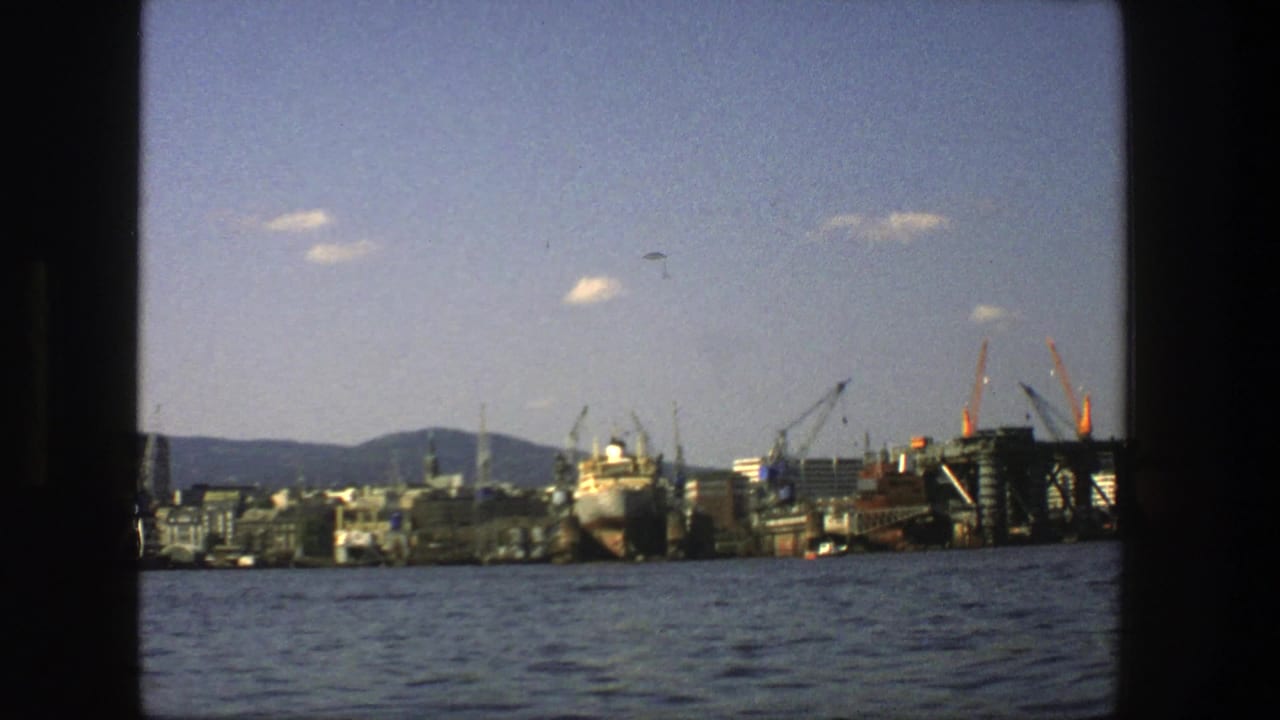 Harbor waterfront with cranes, a large ship, city buildings, blue water, hills, and scattered clouds.