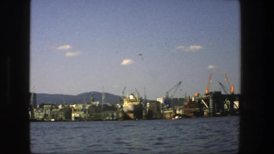 Stockholm seaport in 1975 viewed from the water, showing cranes, a large ship, low city buildings, distant hills, and small clouds.