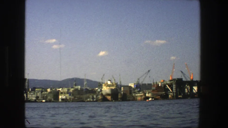 1975 Stockholm waterfront view across the harbor, framed by dark film edges and showing cranes, a ship, buildings, hills, and blue water.