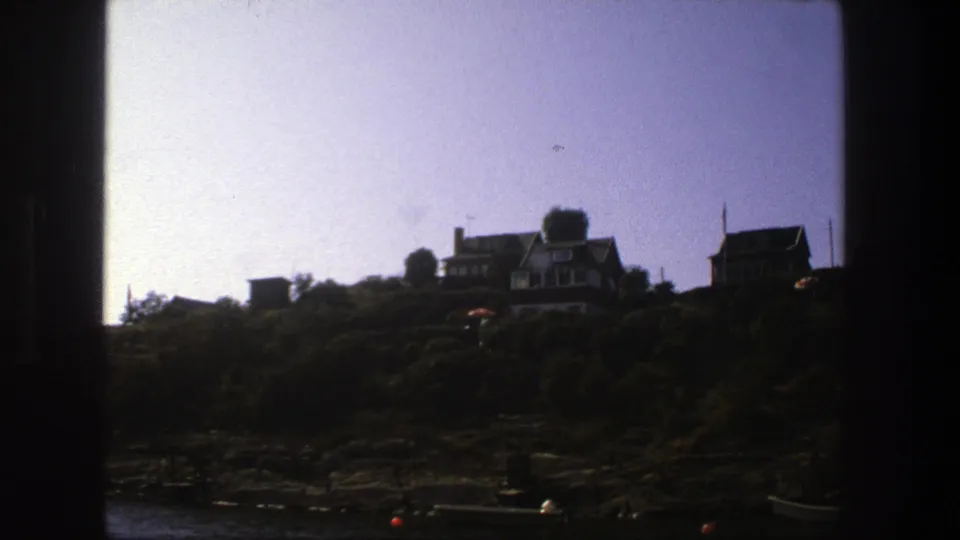 Stockholm shoreline in 1975 with houses silhouetted on a rise above rocks and water, seen through a dark-edged home-movie frame.